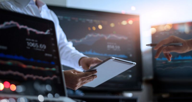 Businessman using tablet for analyzing data stock market in monitoring room with team pointing on the data presented in the chart