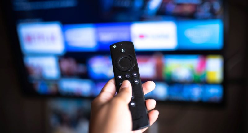 TV remote in hand with selective focus TV background.