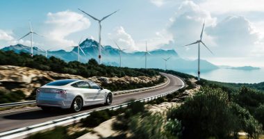 Electric car drives on a highway with wind turbines in background.